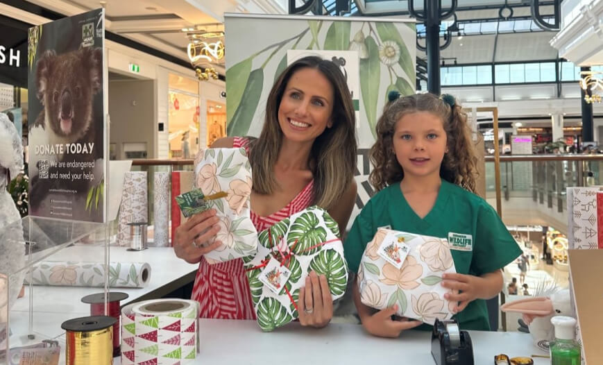 Woman and child holding wrapped gifts at a koala conservation fundraising station in a shopping mall.