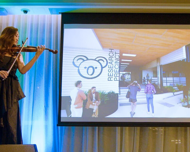 Woman playing violin, presentation screen shows Research Precinct architectural design.