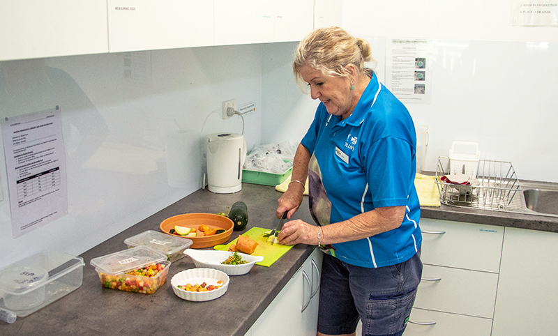 A woman in a blue shirt slices vegetables on a cutting board in a kitchen, with containers of chopped produce on the counter.