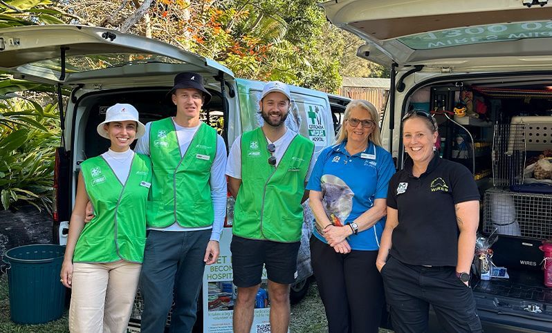 Five people stand in front of open vehicles, with three wearing green vests and two in blue and black uniforms, outdoors near trees and animal carriers.