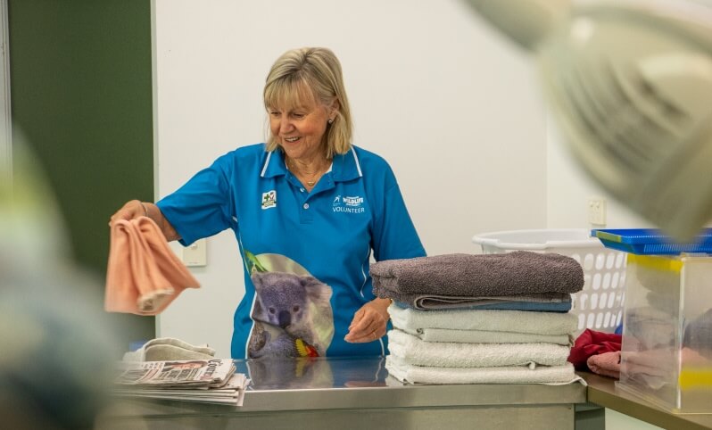 A woman in a blue volunteer shirt folds towels in a laundry area, with a laundry basket and folded towels on the counter.