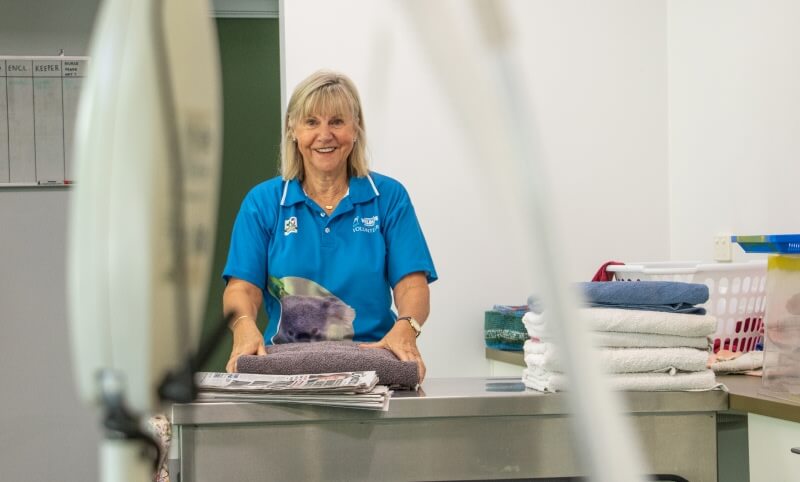 A woman wearing a blue polo shirt stands behind a counter, folding towels in a laundry room with a basket of folded laundry beside her.