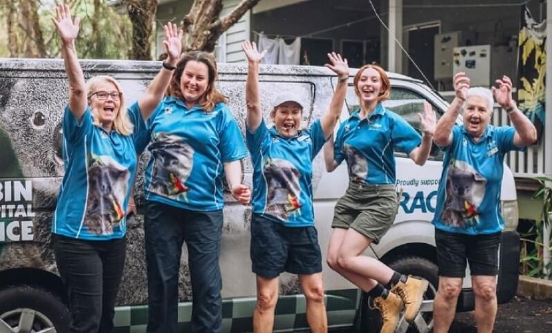 Five women in matching blue shirts stand in front of a Currumbin Wildlife Hospital ambulance van, smiling and posing with raised arms in an outdoor setting.