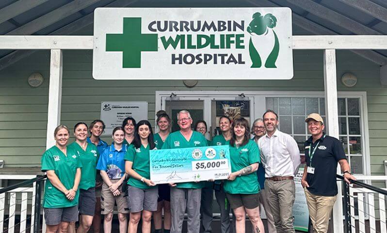 A group of people stands outside Currumbin Wildlife Hospital holding a large $5,000 donation check in front of the hospital’s entrance.