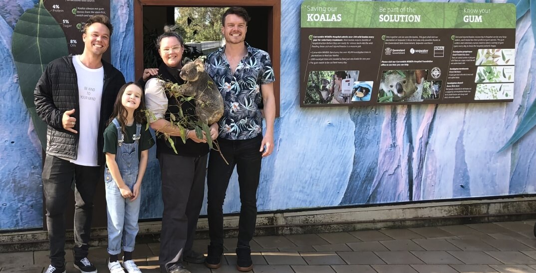 Four people, including a child, pose with a koala held by one person in front of an informational display about koalas at a zoo or sanctuary.