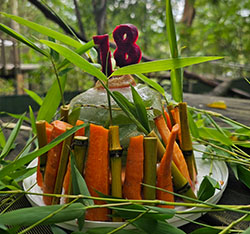 A dome-shaped vegetable arrangement with carrots and bamboo on a plate, topped with a purple 18 decoration and surrounded by green leaves.