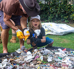 An adult and a child wearing gloves collect and sort litter outdoors, with a pile of trash on the ground in front of them.