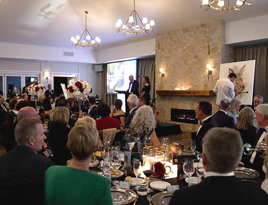 A formal dinner event with seated guests facing a stage where two people are speaking at a podium, with a projected screen and fireplace in the background.