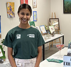 A young woman stands next to a table with brochures, a sign reading Art Exhibition Fundraiser, and art displayed in the background.