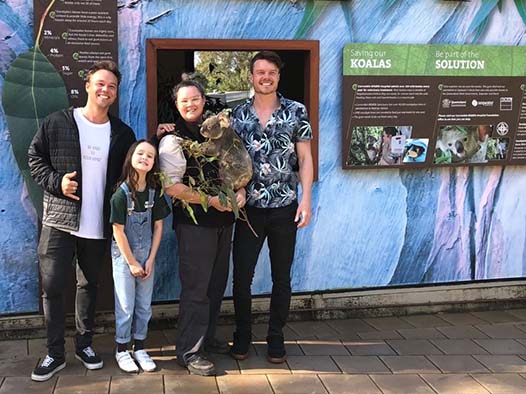 Four people pose in front of informational zoo signs; one person holds a koala. They stand on a paved walkway outdoors.