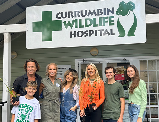 A group of seven people stand in front of the Currumbin Wildlife Hospital entrance, smiling at the camera. The hospital sign features a green cross and a koala logo.