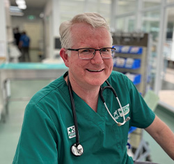A smiling medical professional in green scrubs and a stethoscope sits in a clinical setting with medical equipment in the background.