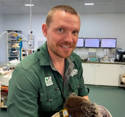 A man in a green uniform stands in a medical or veterinary clinic, holding an animal. Medical equipment and monitors are visible in the background.
