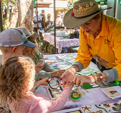 A woman in a yellow shirt and hat helps children examine small objects with magnifying glasses at an outdoor table covered with educational materials.