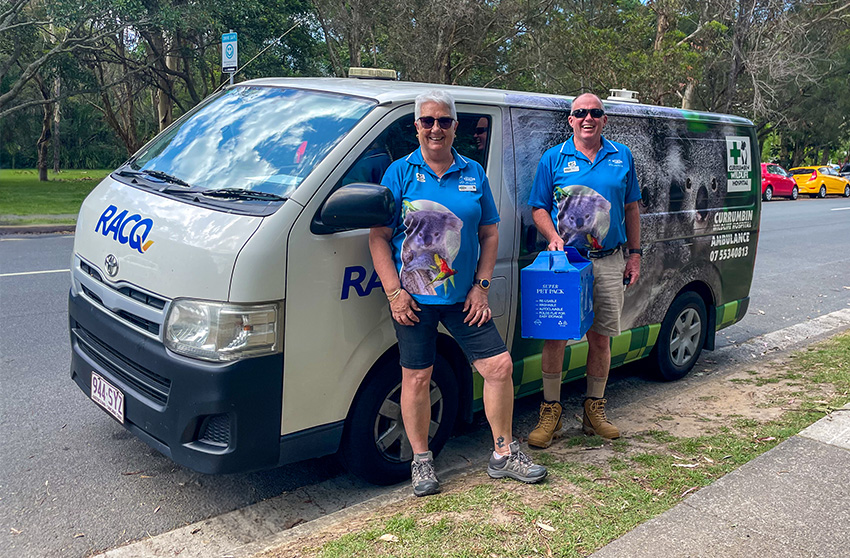 Two people in blue wildlife shirts stand beside a RSPCA van parked on a roadside, with trees and cars in the background.