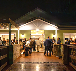 People are gathered outside and inside a well-lit building labeled Homestead at night, socializing on a wooden deck and near the entrance.