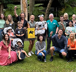 A group of people of various ages pose outdoors on grass, some standing and some sitting, holding a koala sign and a koala stuffed animal.