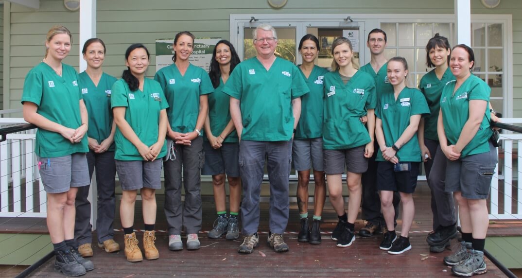 Twelve people wearing green scrubs and grey shorts stand in two rows on a wooden porch in front of a building, smiling at the camera.