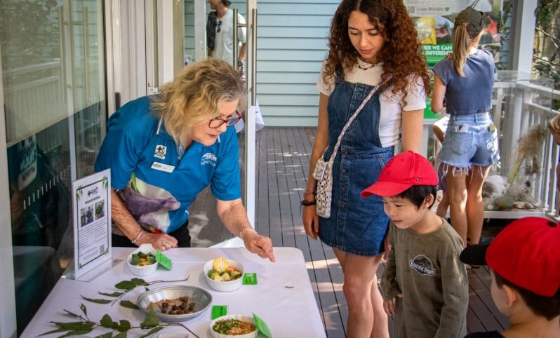 An older woman explains samples of food to a young woman and two children at an indoor event table.