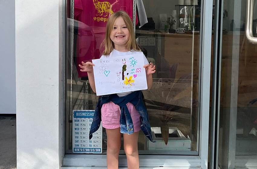 A young girl stands in front of a glass door, smiling and holding up a colorful drawing. Store hours are displayed on a sign next to her.