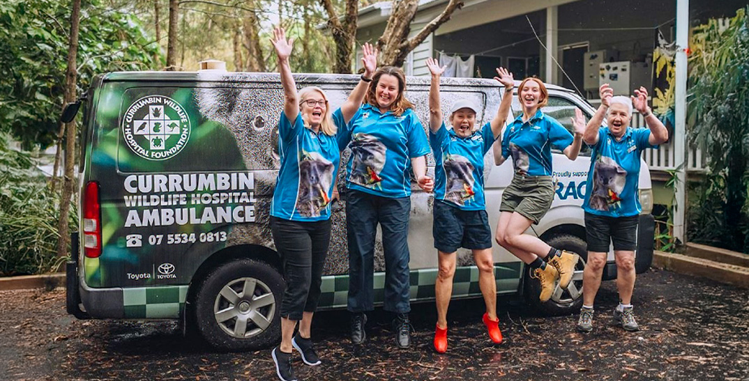 Five women in matching blue shirts stand in front of a Currumbin Wildlife Hospital ambulance van, smiling and posing with raised arms in an outdoor setting.