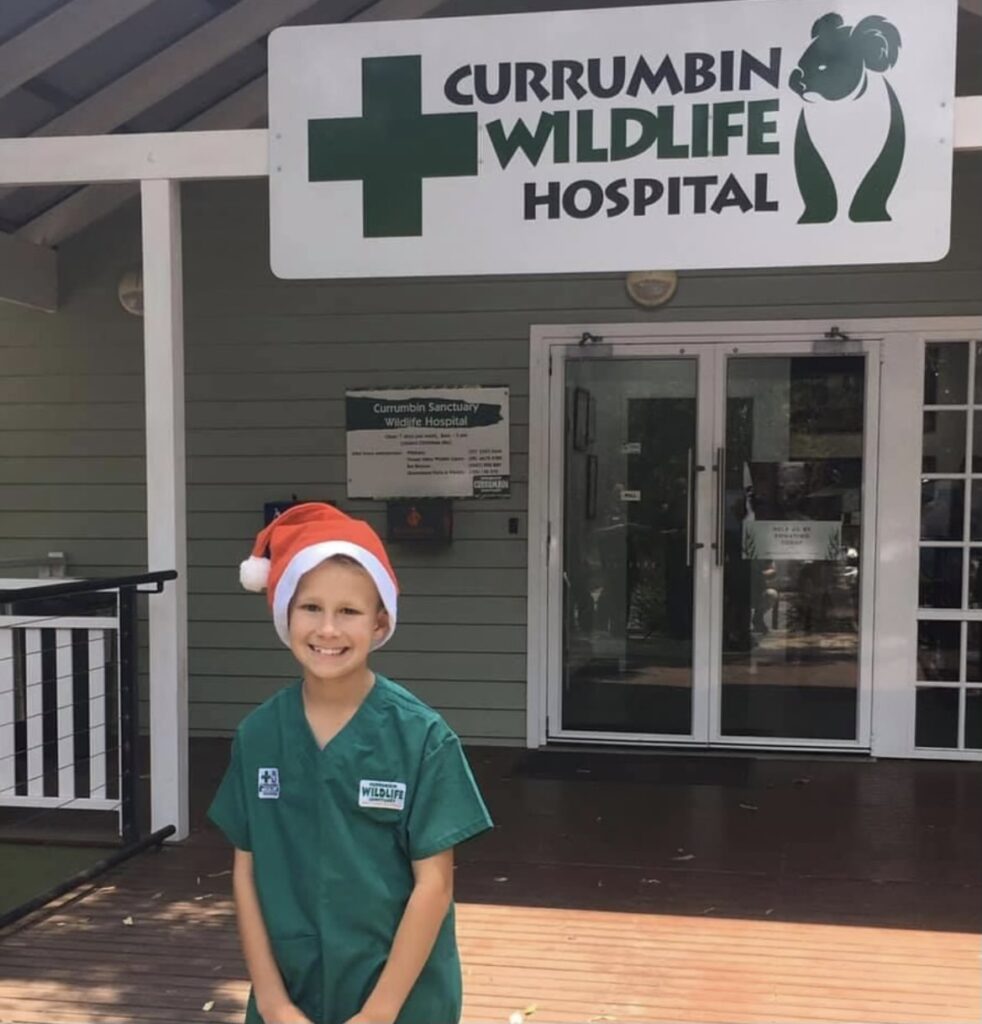 Young smiling boy wearing a green clinical shirt and Christmas hat, standing in front of Currumbin Wildlife Hospital