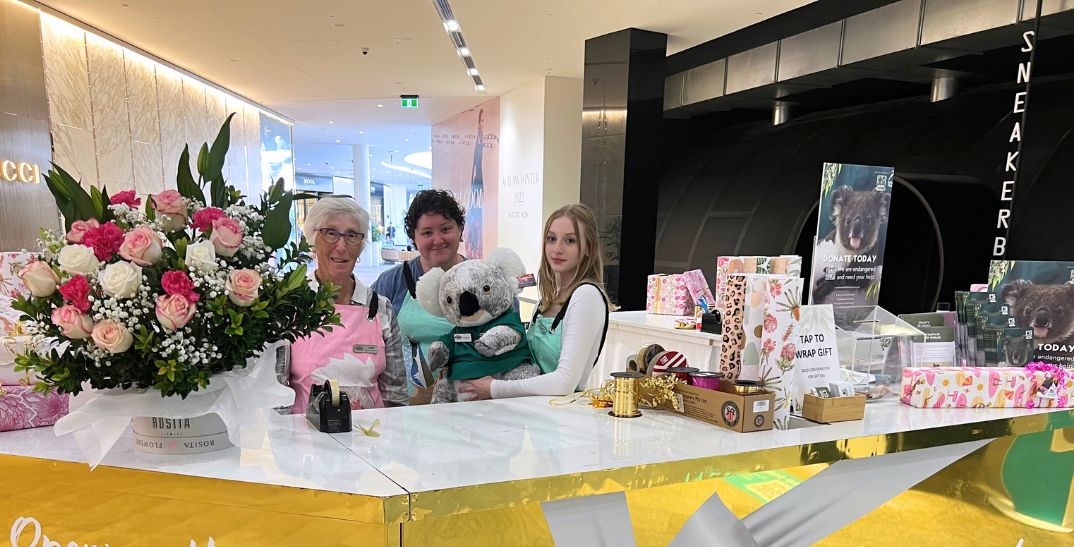 Three women behind a gift wrapping stand wrapped in gold and silver paper. One of the women is holding a large Koala plush. On the stand is a large bouquet of roses and beautifully wrapped gifts