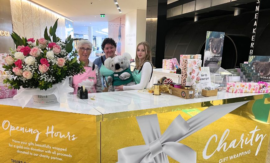 Three women behind a gift wrapping stand wrapped in gold and silver paper. One of the women is holding a large Koala plush. On the stand is a large bouquet of roses and beautifully wrapped gifts