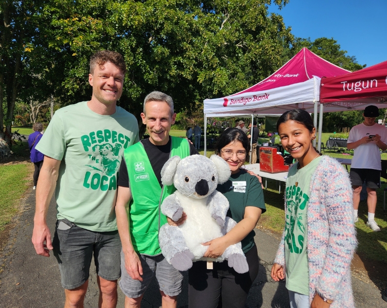 A group photo of two women and two guys, all smiling wearing green tops with print respect locals. One of the women is holding up a large plush Koala. Behind them are stalls, people and large trees