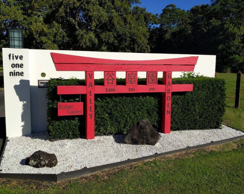 A Japanese style red sign saying Tani Tei En Valley Garden, sitting on a bed of pebbles in front of a manicured hedge and white stone wall