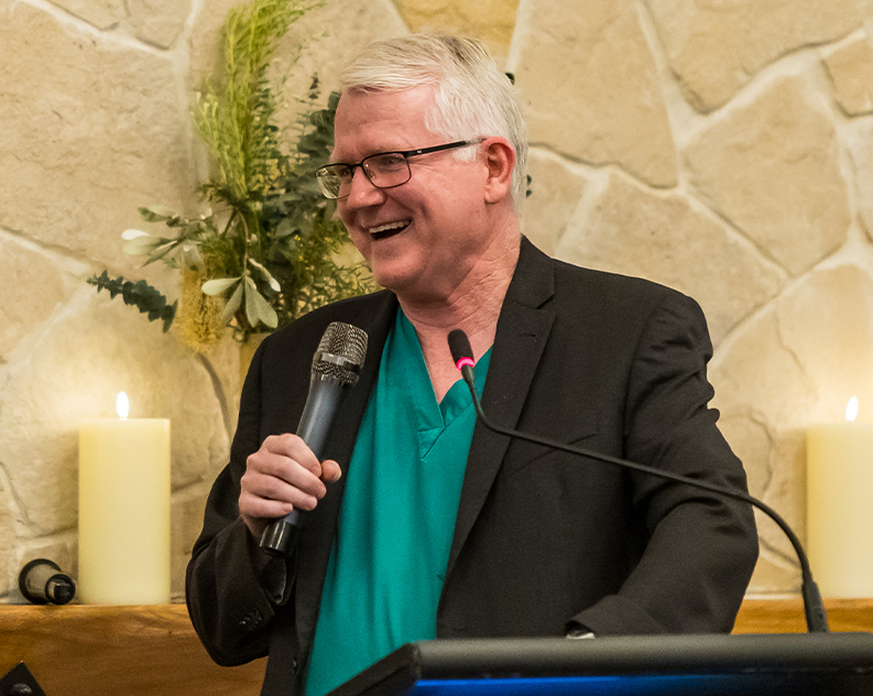 A man wearing a teal veterinarian shirt underneath a black blazer holding a microphone, smiling while standing in front of a lectern. He is standing in front of a neutral rock wall with a wall shelf holding large burning candles and native flower bouquets