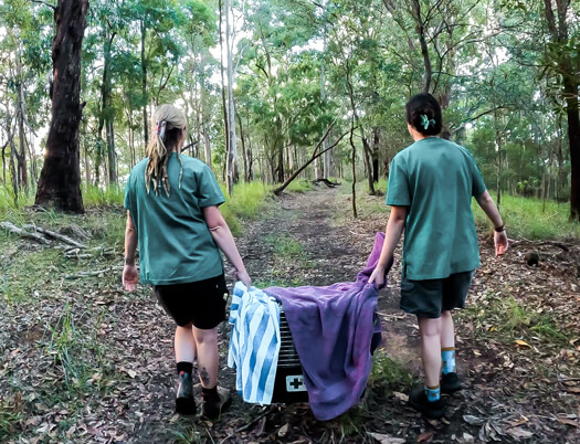 Two people in green shirts carrying a pet carrier in a forest.