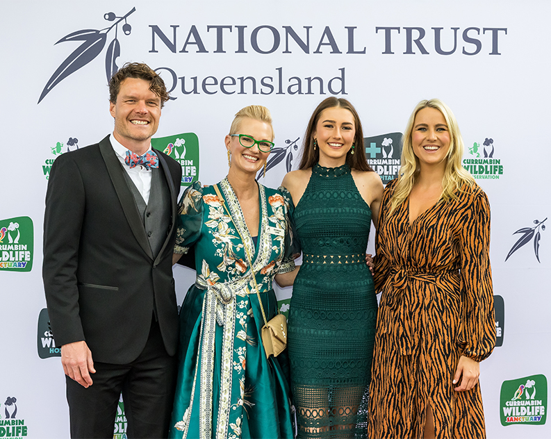 Three women in cocktail dresses and a man in a black suit posing for camera in front of a white photo wall with several logos printed on it