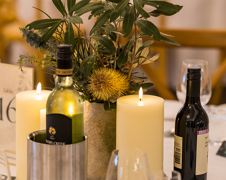 A table set a white table cloth topped wtih bottles of wine, large burning candles and a bouquet of Australian native plants