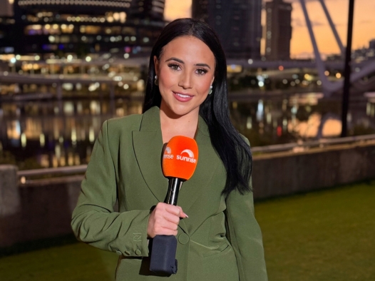 A female reporter smiling to the camera holding a bright orange microphone. She is wearing a navy green suit and appears to stand in front of the Brisbane river at dusk, with the sunset and lights lighting up the buildings and sky in orange hues