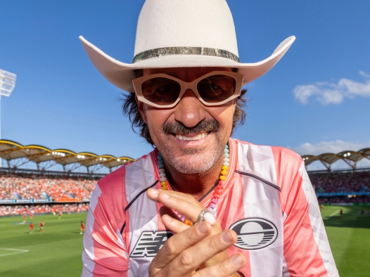 A guy wearing a white cowboy hat and funky classes closeup smiling and putting his hands together. He is in an open-air Australian football stadium on a sunny day with clear blue skies