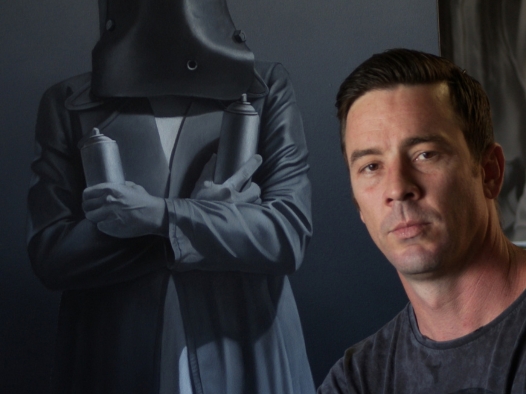 A man's headshot wearing a dark grey t-shirt looking seriously at the camera. Right behind him is a dark clad figure holding dark grey candles and wearing a black face cover.