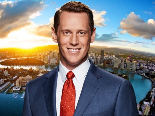 A male reporter's headshot as he is looking straight on and smiling, wearing an elegant white shirt and suit jacket with a bright red tie. He appears on an image background of Brisbane on a partly cloudy day with the sun rising