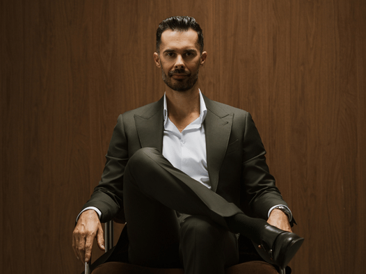Real estate legend and Gold Coast personality Billy Jaz sits in a seat in a nice suit with his legs crossed staring into the camera